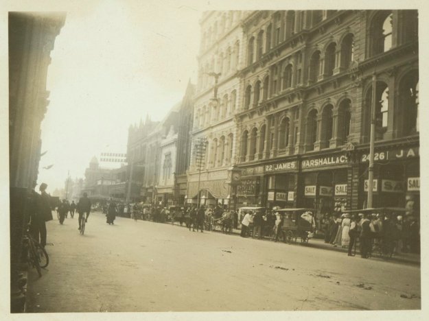View looking down a street, with horse-drawn vehicles parked along the curb and pedestrians walking along the footpath. A sign on a building reads: 22. James Marshall & Co. 24-26.